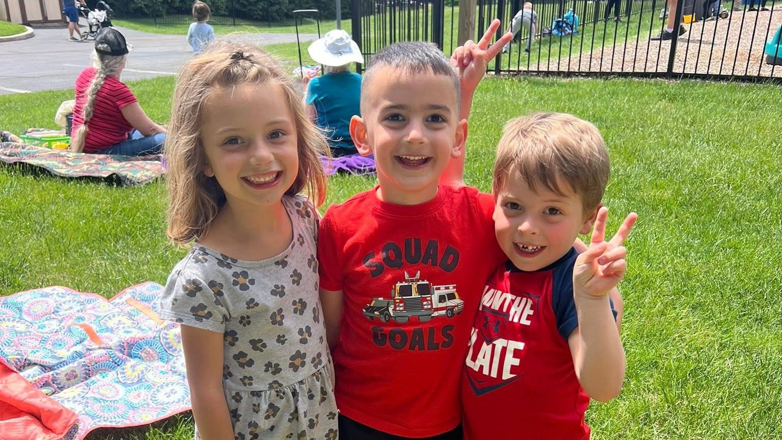 Three smiling Open Arms friends arm in arm on the lawn at the school picnic
